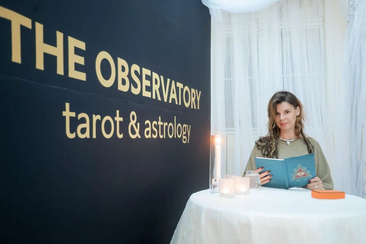 Rezi at a table with cards and candles in front of a black wall reading The Observatory — tarot & astrology